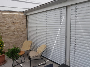 Zusehen ist ein Balkon mit einer Steinwand, zwei Stühlen mit Fellbezug und zwei Pflanzen. Dahinter sind silberne geschlossene Lamellenstoren.